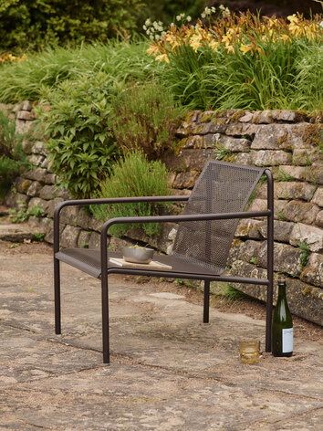 Dapple Loungestol i färgen Dark Chocolate som står på en stenig uteplats fylld med blommor och grönska